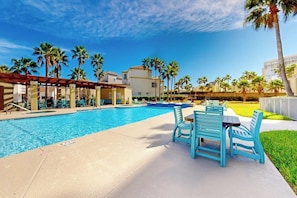 A swimming pool with lounge chairs and a covered seating area, surrounded by palm trees and buildings. - Palm Frond Sanctuary, Unit 302 (South Padre Island)