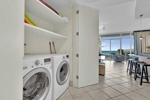 A modern laundry room with two front-loading washing machines, a shelf with cleaning supplies, and a view of a balcony with outdoor seating. - Sapphire Condominiums #408 (South Padre Island)
