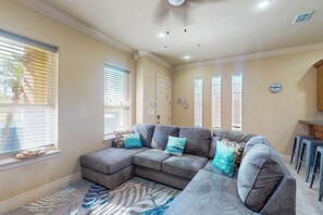 A living room with a sectional sofa, a patterned rug, and a kitchen area with stools. - Island Breeze Retreat on Jupiter (South Padre Island)