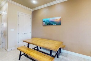 Une pièce avec une table et des bancs en bois, des murs de couleur beige et un tableau représentant un coucher de soleil sur l’océan.