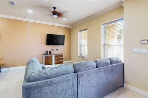 A living room with a sectional sofa, a flat-screen TV mounted on the wall, a ceiling fan, and a window with blinds. - Island Breeze Retreat on Jupiter (South Padre Island)