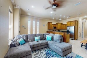 A living room with a sectional sofa, a ceiling fan, and a kitchen area with wooden cabinets and a refrigerator. - Island Breeze Retreat on Jupiter (South Padre Island)