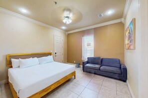 A bedroom with a bed, a gray sofa, a window with blinds, and a ceiling fan. - Island Breeze Retreat on Jupiter (South Padre Island)