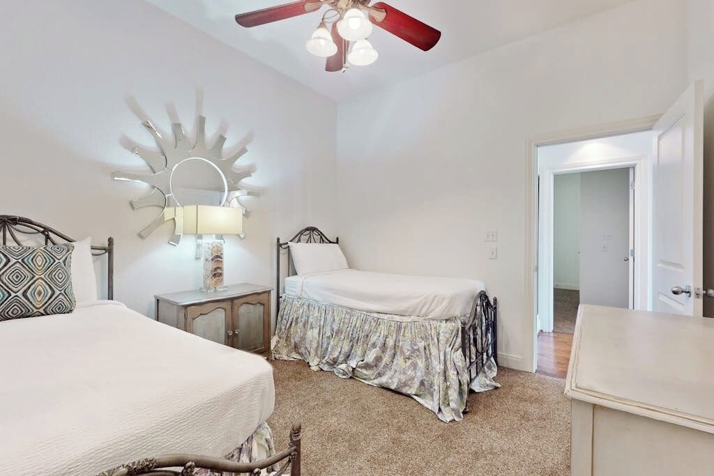 Un dormitorio con cama, mesitas de noche, lámpara, ventilador de techo y puerta que da a otra habitación.