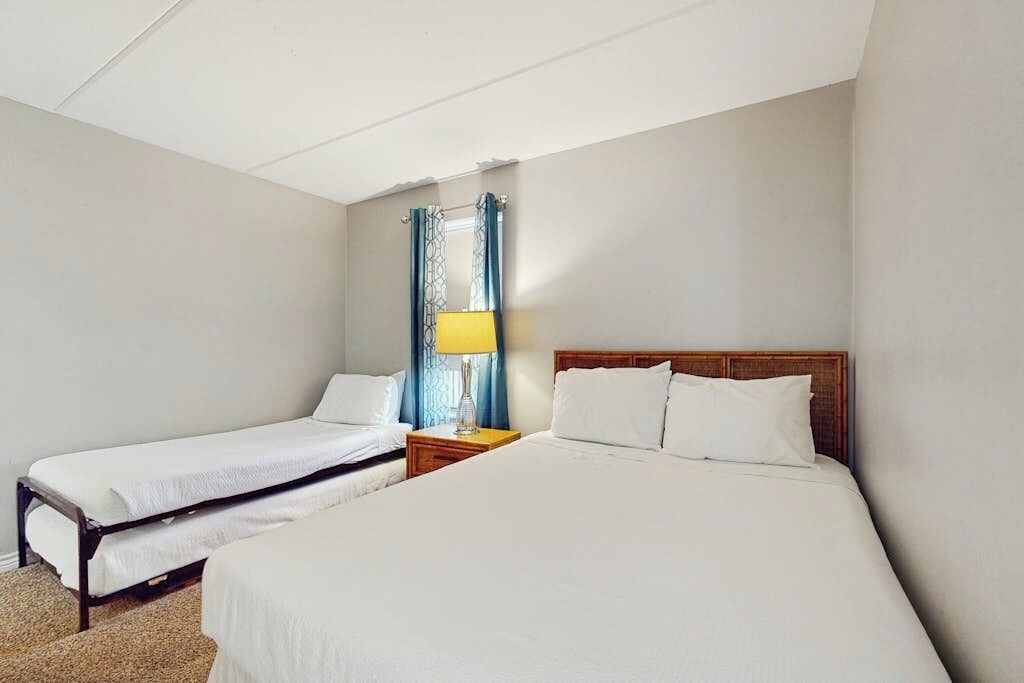 Habitación de hotel con dos camas individuales, cabecero de madera, mesita de noche con lámpara y ventana con cortinas.