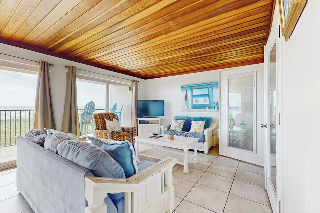 A spacious living area with a wooden ceiling, a sofa, a television, and a balcony with a view.