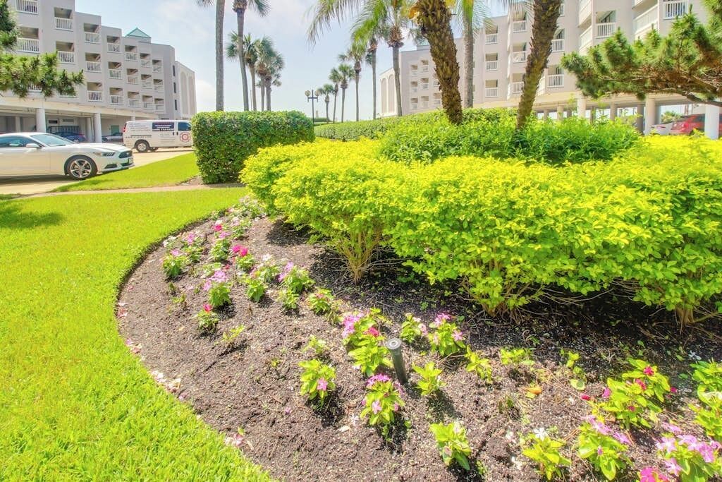 Un espace paysager aménagé en jardin, avec une variété de plantes et d’arbustes, un chemin pavé et, au fond, un bâtiment doté de plusieurs balcons.