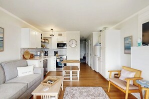 A modern kitchen with white cabinets, a dining area with a wooden table and benches, and a living area with a sofa and a chair. - Wandless Waves - Pool & Hot Tub, Steps to Sand (Galveston)
