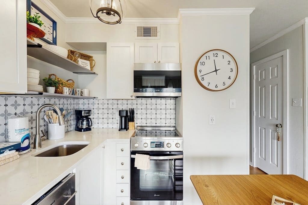 Een keuken met witte kastjes, een roestvrijstalen oven, een wandklok en een houten eettafel.