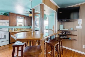 A kitchen with wooden cabinets, a central island with stools, a built-in oven, and a television mounted on the wall. - Down By The Sea - Private Pool, Downtown Port A (Port Aransas)
