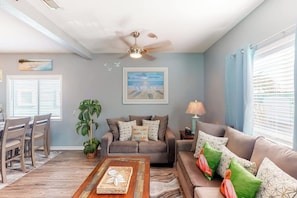 A living room with a ceiling fan, a dining area with chairs, a sofa with cushions, a coffee table, and a painting on the wall. - Leeward Isles 305 - Pool, Near the Beach, Dogs OK (Corpus Christi)