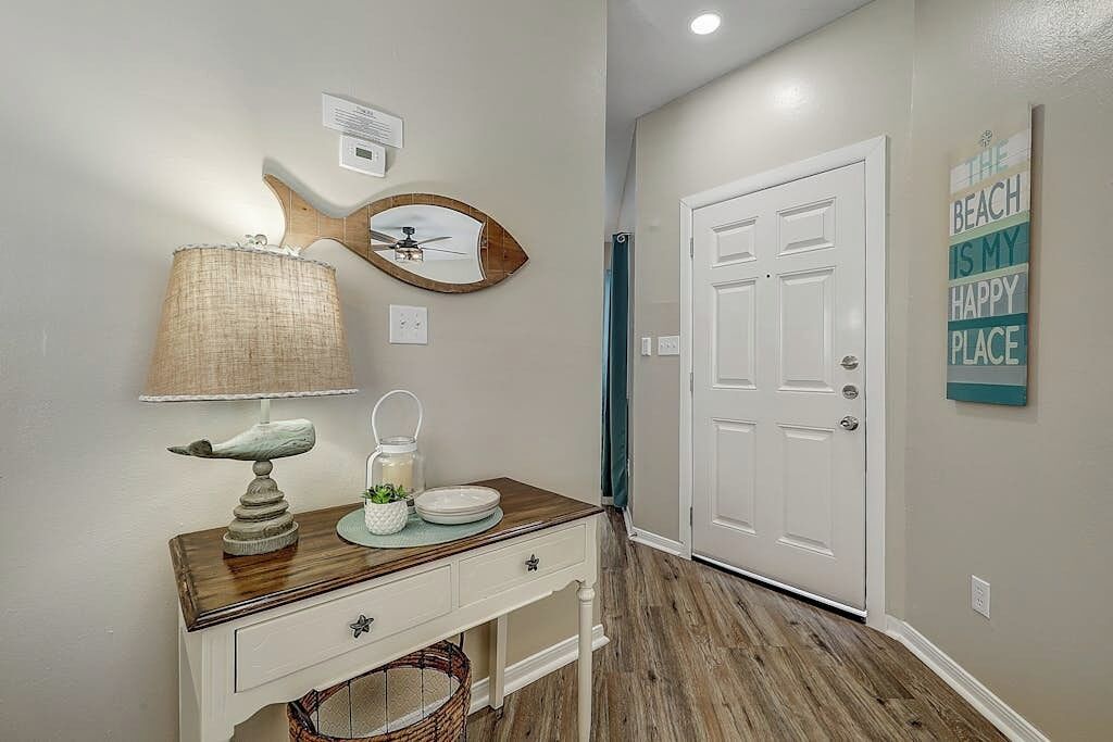 Un couloir avec un plancher en bois, une porte blanche, un miroir fixé au mur, une lampe et une assiette décorative.