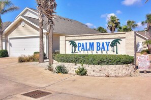 A sign for Palm Bay Village with a garage and palm trees. - Palm Bay 307 - Heated Pool, Walk to the Beach (Corpus Christi)