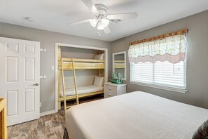 A bedroom with a bunk bed, a single bed, a ceiling fan, a window with curtains, and a bedside table. - The Moor The Merrier (Port Aransas)