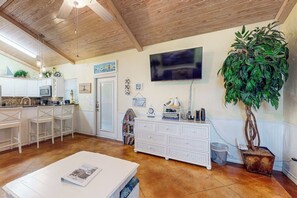 Eine Küche mit weißen Schränken, ein Essbereich mit Tisch, ein an der Wand befestigter Fernseher und ein Blumentopf.
