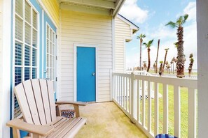 A porch with a blue door, a wooden chair, and a white railing overlooking palm trees. - Beach Club 173 - Pool, Sauna & Beach Access (Corpus Christi)