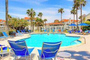 A swimming pool with lounge chairs and umbrellas, surrounded by palm trees and buildings. - Beach Club 173 - Pool, Sauna & Beach Access (Corpus Christi)