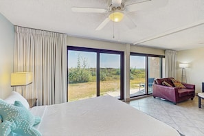 A hotel room with a bed, a red sofa, a standing lamp, and a view through large glass doors. - Island House #105 - Heated Pool & Outdoor Patio (Corpus Christi)