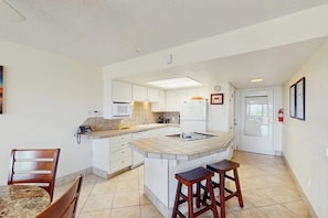 A compact kitchen with a central island, white cabinetry, and a dining area with a marble table and wooden chairs. - Island House #105 - Heated Pool & Outdoor Patio (Corpus Christi)