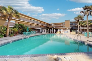 A swimming pool with a clear blue water, surrounded by a multi-story building with balconies, palm trees, and outdoor seating. - Island House #105 - Heated Pool & Outdoor Patio (Corpus Christi)