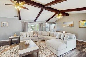 Ein geräumiges Wohnzimmer mit einem großen Ecksofa, Holzbalken, Deckenventilatoren und Blick durch das Fenster.