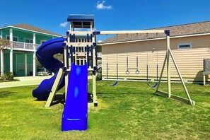 Um parquinho infantil com um escorrega azul e balanços em um bairro residencial.