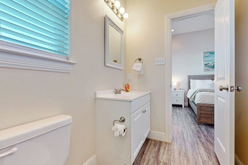 Un bagno con il water, un lavandino e uno specchio. Si apre una porta su una camera da letto con un letto e un comodino.