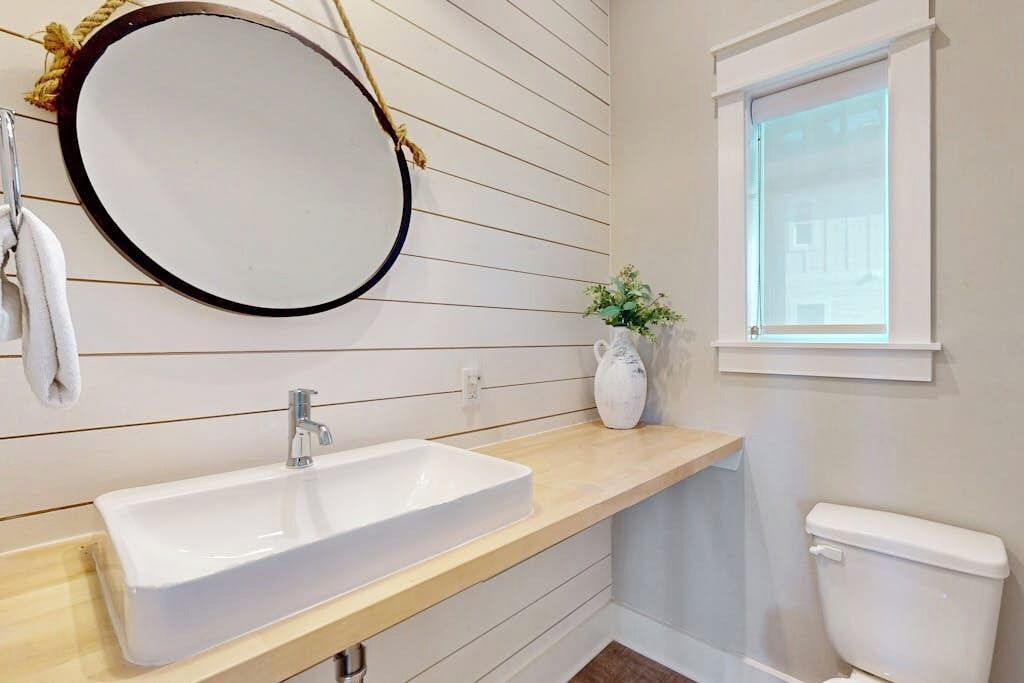 Ein Badezimmer mit einem weißen Waschbecken, einem runden Spiegel, einem Fenster und einer Toilette.