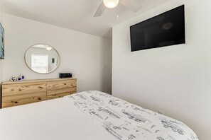 A bedroom with a bed, a wooden dresser, a round mirror, a flat-screen TV mounted on the wall, and a ceiling fan. - CopaCabana - Pool & Patio, Near the Beach (Port Aransas)