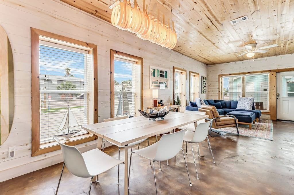 A spacious living area with a wooden ceiling, a dining table with chairs, a blue sofa, and large windows with blinds.