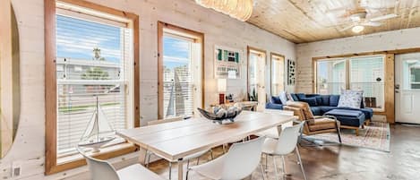 A spacious living area with a wooden ceiling, a dining table with chairs, a blue sofa, and large windows with blinds.