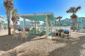 A covered outdoor seating area with picnic tables and chairs, surrounded by palm trees and a building with a light blue facade. - CopaCabana - Pool & Patio, Near the Beach (Port Aransas)