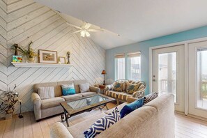 Ein Wohnzimmer mit einer Wand im Fischgrätmuster, einem Deckenventilator, einer Sofagarnitur, einem Couchtisch und Blick durch eine Glastür.