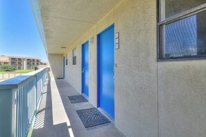 A corridor with blue doors, a balcony with a railing, and a building in the background. - Ocean Wave - Pool, Walk to Beach & Town (Port Aransas)