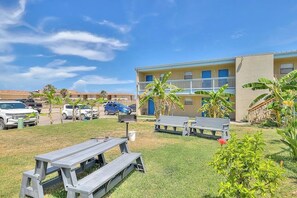 A courtyard with picnic tables, a building with a balcony, and parked cars. - Ocean Wave - Pool, Walk to Beach & Town (Port Aransas)