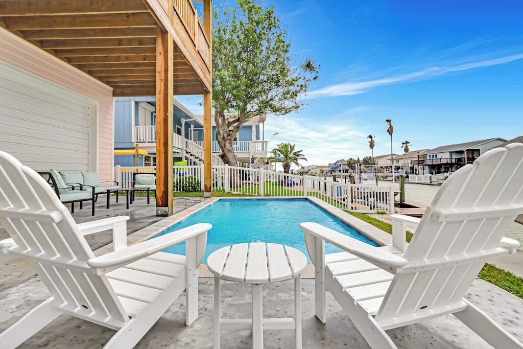 A pool area with white Adirondack chairs and a small table, surrounded by a white fence and a tree.