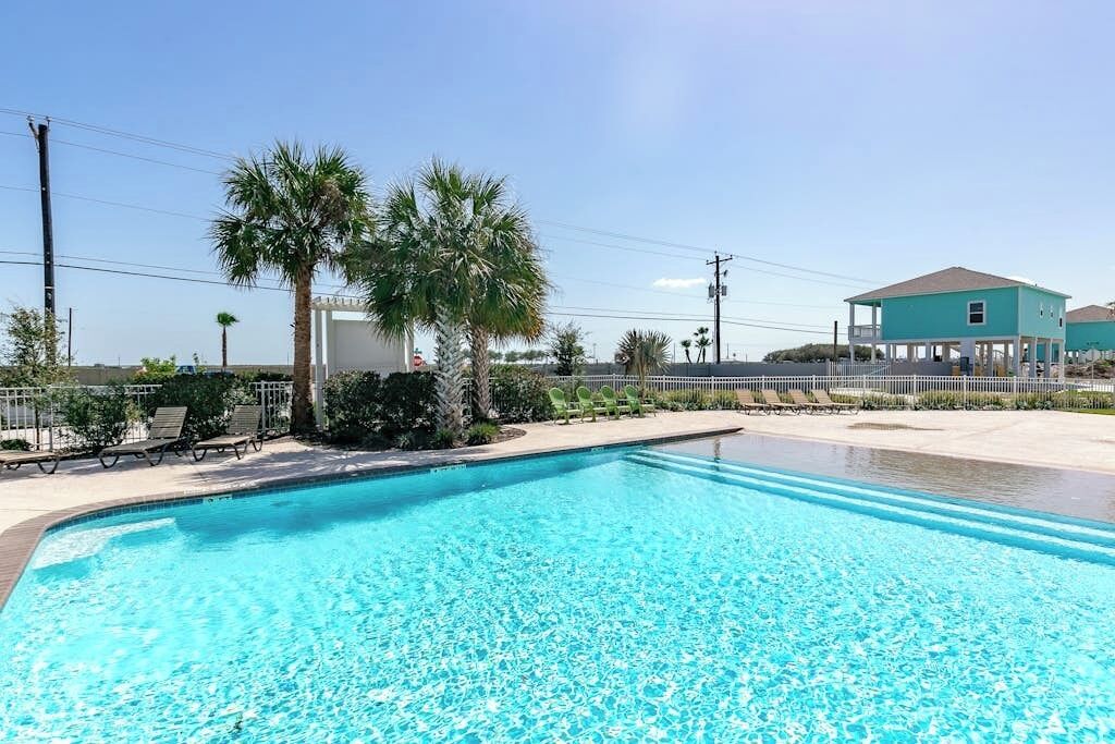 A swimming pool with clear blue water, surrounded by palm trees and a fenced area.