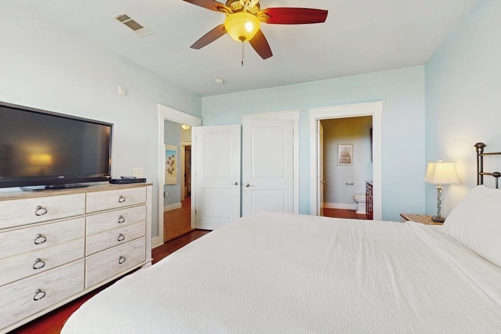 A hotel room with a bed, a television on a dresser, a ceiling fan, and a door leading to another room.