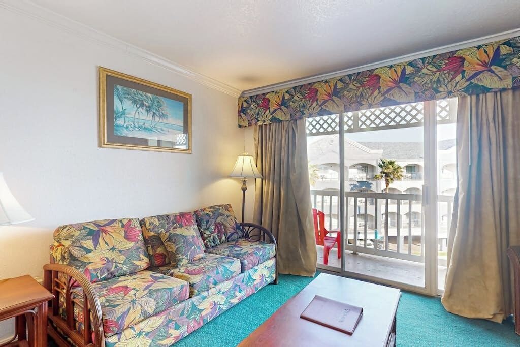 Een kamer met een bank met bloemenprint, een houten bijzettafeltje, een balkon met uitzicht op gebouwen en een rode stoel.