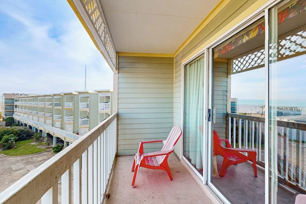 A balcony with two red chairs, a view of a building with multiple balconies, and a clear sky.