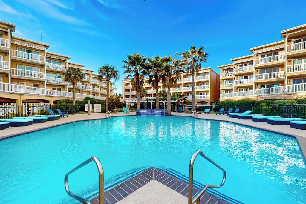 A hotel pool area with multiple balconies, palm trees, and lounge chairs.