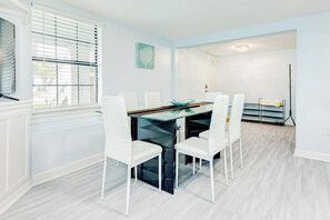 A dining area with a glass-top table, white chairs, and a view of a room with a bed and a window. - Ocean Waves - Hot Tub, Fenced Yard & Dogs OK (Galveston)