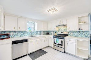A kitchen with white cabinets, a tiled backsplash, a dishwasher, a stove, and a microwave. - Ocean Waves - Hot Tub, Fenced Yard & Dogs OK (Galveston)