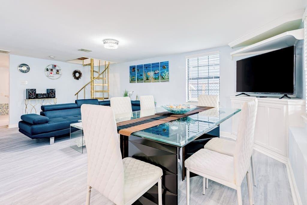 Een moderne woonkamer met een glazen eettafel, witte stoelen, een blauwe bank en een aan de muur bevestigde televisie.