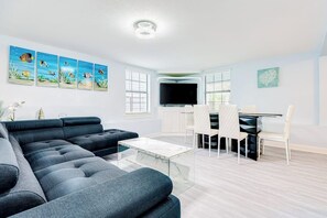 A modern living room with a sectional sofa, a dining table with chairs, a flat-screen TV, and artwork on the walls. - Ocean Waves - Hot Tub, Fenced Yard & Dogs OK (Galveston)