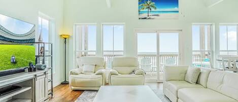 A spacious living room with a large flat-screen TV, two white leather sofas, a coffee table, and a view of the ocean through large windows.