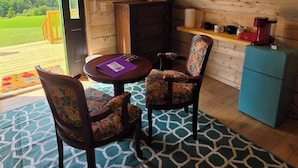 A cozy room with a wooden ceiling, a round table with chairs, a small cabinet, and a view of a green landscape through a door.