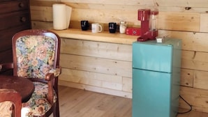 A wooden cabin interior with a wooden dresser, a floral chair, a teal refrigerator, and a shelf with mugs and a coffee maker.