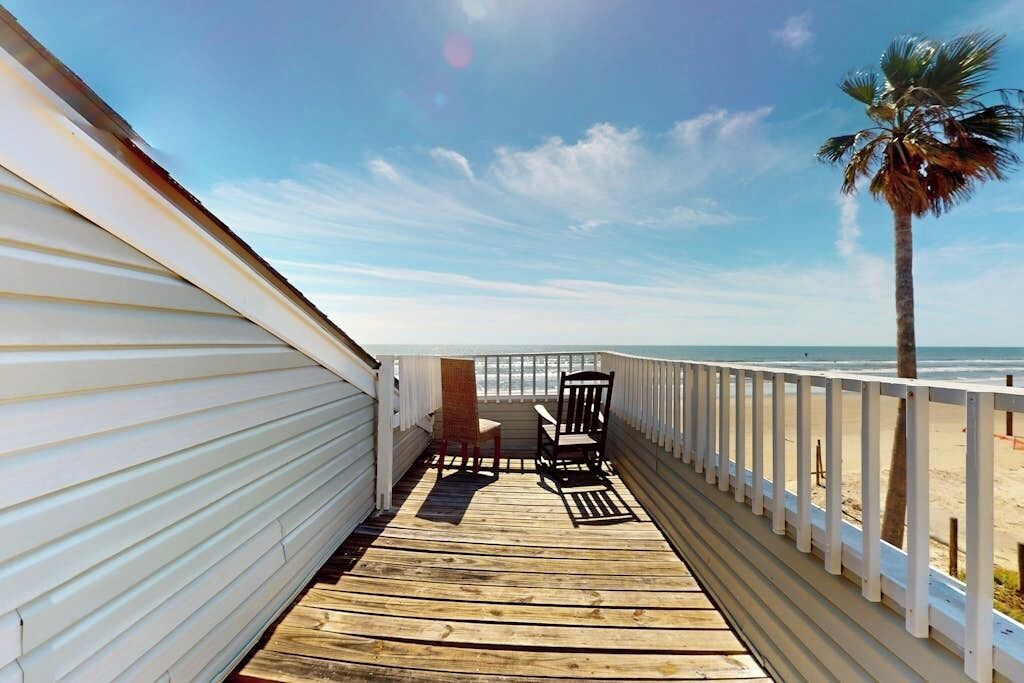 Una terraza de madera con barandilla, una palmera y vista a la playa.