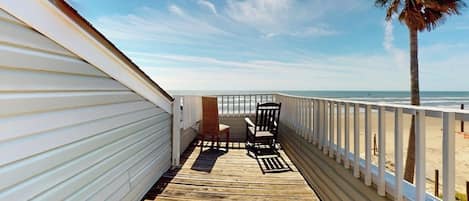 Una terraza de madera con barandilla, una palmera y vista a la playa.